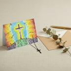 A colorful religious art print or card featuring a crucifix scene, shown standing on a white background. The artwork depicts a glowing cross with a bright blue and yellow center, fiery orange and red sky, green ground.