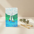 A rectangular greeting card, featuring a colourful illustration of a sailboat on blue water with green hills and white clouds. The left side of the card is plain white with the AllThingsGoode logo.