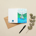 A rectangular greeting card, featuring a colourful illustration of a sailboat on blue water with green hills and white clouds. The left side of the card is plain white with the AllThingsGoode logo.