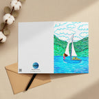 A rectangular greeting card, featuring a colourful illustration of a sailboat on blue water with green hills and white clouds. The left side of the card is plain white with the AllThingsGoode logo.